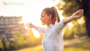 A young woman standing outdoors with her arms outstretched and eyes closed, smiling peacefully in the sunlight, surrounded by blurred greenery and a building in the background; represents joy, freedom, and spiritual renewal.