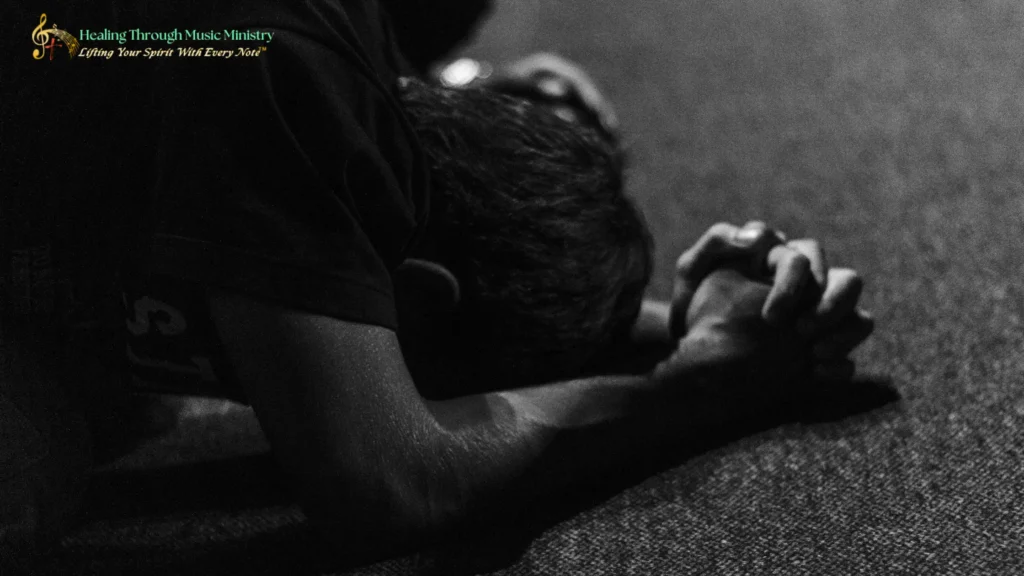 A person kneeling on the floor with head bowed and hands clasped in prayer, shown in black-and-white lighting, conveying a quiet and reflective mood.