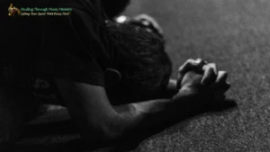 A person kneeling on the floor with head bowed and hands clasped in prayer, shown in black-and-white lighting, conveying a quiet and reflective mood.