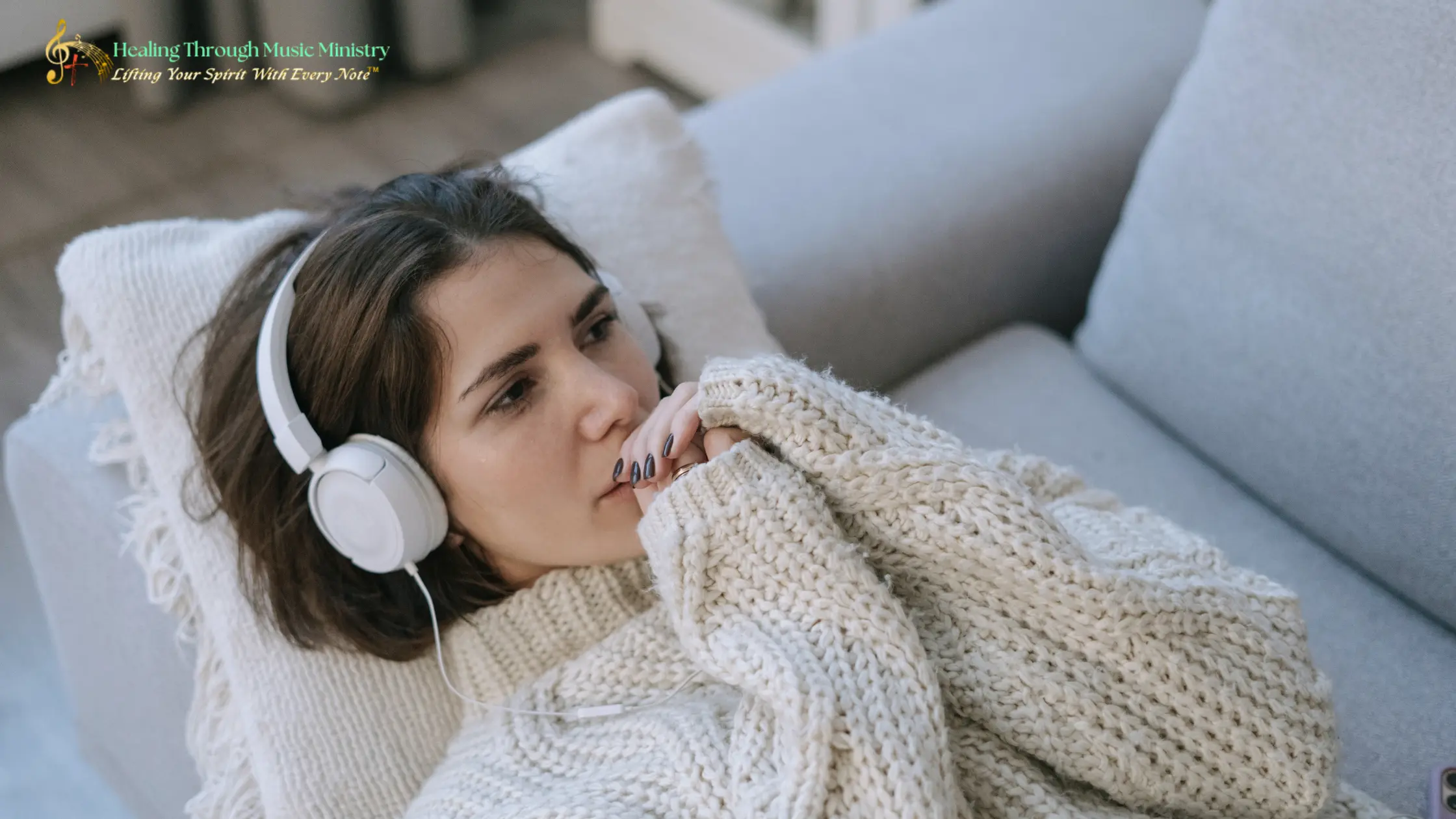 A young woman wearing white headphones lies on a light-colored couch, wrapped in a chunky cream knit sweater. She gazes thoughtfully into the distance while listening to audio, creating a calm and reflective mood.